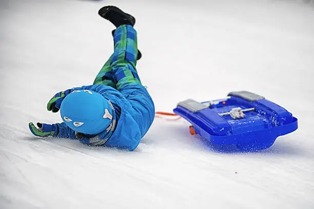 Little boy riding toboggan or sled a bit too fast. Cold winter day. The boy has fallen off toboggan and is sliding down hill.