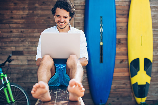 Cheerful young man using laptop with surfing boards at the background.