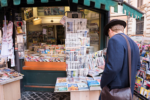 Rome, Italy - April 15, 2016: A customer (male wearing a hat and carrying a bag over his shoulder) is looking for a newspaper or a magazine at a traditional newsstand near Piazza della Rotonda in Rome, Italy, on a morning in spring. The newsstand offer both Italian as well as international newspaper including Die Welt, Le Monde, International New York Times, Financial Times, Daily Mail, El Pais and The Guardian - as well as magazines and books (and probably snacks, drinks and souvenirs).