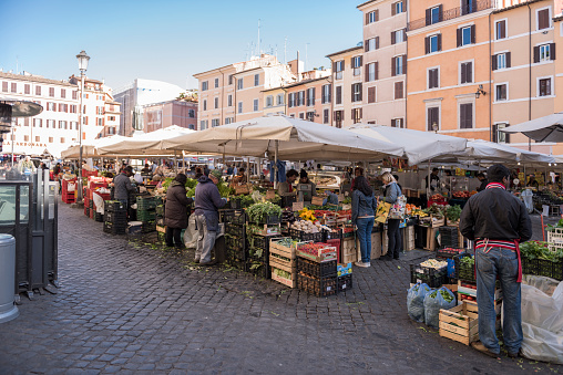 Rome, Italy - April 15, 2016: The street market of Campo de Fiori in the center of Rome in Italy on a morning in spring. The vendors in the nearest stalls are offering fruit and vegetables. Some customers are buying food. Other types of raw and processed food are also offered, as well as clothes and souvenirs. Shallow depth of field. Focus was placed over the nearest market stalls, vendors and customers. The background is blurred.