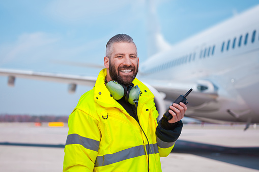 Ground crew using walkie-talkie outdoor in front of aircraft at the airport, smiling at camera.
