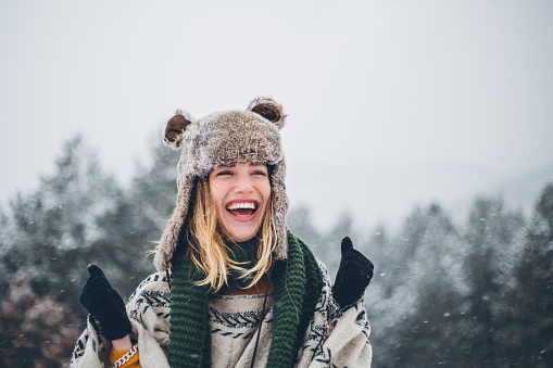 Outdoor close up portrait of young beautiful happy smiling hipster girl, wearing stylish poncho and fake fur hat. Model expressing joy and looking at camera. Magic snowfall effect.
