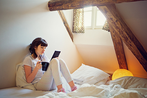 Young teenage girl is reading ebook sitting on the bed in the loft. 