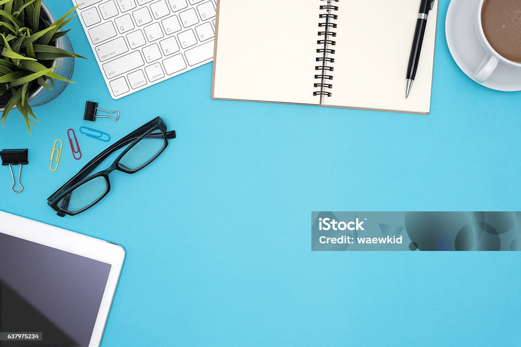 Office desk table with supplies and computer on blue background Office table desk with computer, tablet, pen, coffee cup, flower, eye glasses, supplies and white blank note pad on blue background, Top view and copy space for text Desk Stock Photo