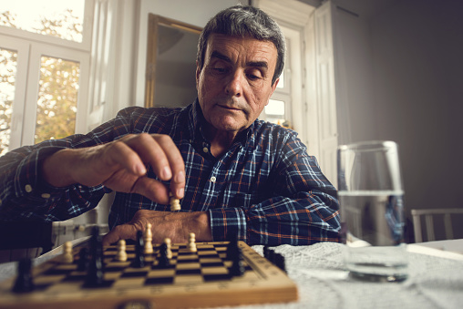 A senior bearded man is playing a chess game at home with his wife. Close-up view of an older man who is making a chess move with a chess piece.