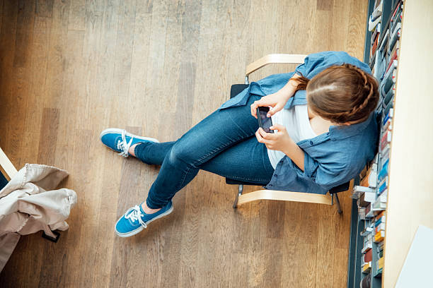 student using smart phone while sitting against bookshelf in classroom Directly above shot of student using smart phone while sitting against bookshelf in classroom woman sitting cross legged in chair stock pictures, royalty-free photos & images