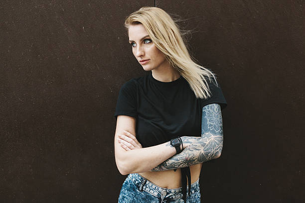 Portrait of a Blonde Tattooed Girl This tattooed woman in Barcelona, Catalonia is not afraid to show her personality and individuality. She is standing next to a wall just as she is - long blonde hair, short blue jeans and a black tee exposing her tattoos. She has a white fixie (fixed gear bicycle) which she uses to cruise the streets right next to her. Behind her is a dark grey wall she is leaning on. black tattoo stock pictures, royalty-free photos & images