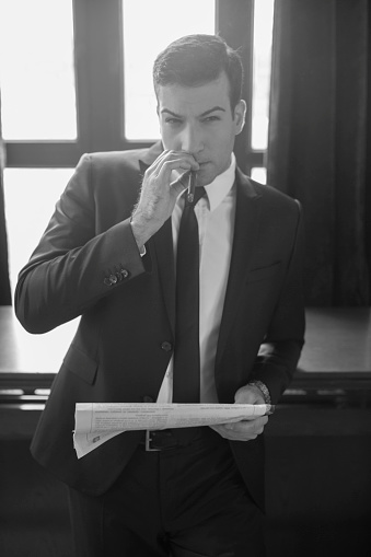 Young handsome businessman standing and leaning leisurely on the table at the cafe, holding newspapers and smoking a cigar