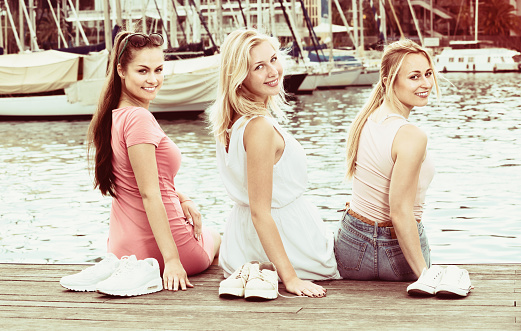 Three cheerful girls sitting together on pier in town on summer day