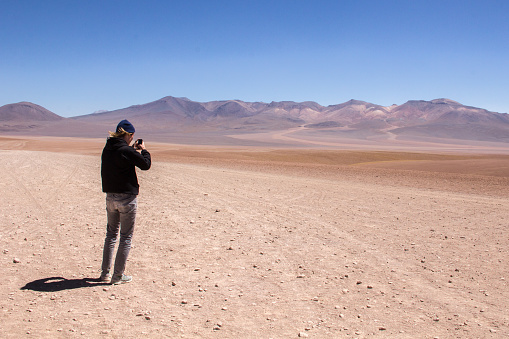 Potosi, Bolivia - October 31, 2016: Photograph of a guy, wearing a black hoodie and grey pants, taking a photograph , with his cellphone, of the dry mountains of a desert