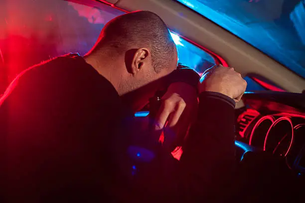 Upset drunk driver is caught driving under alcohol influence. Man covering his face from police car light.