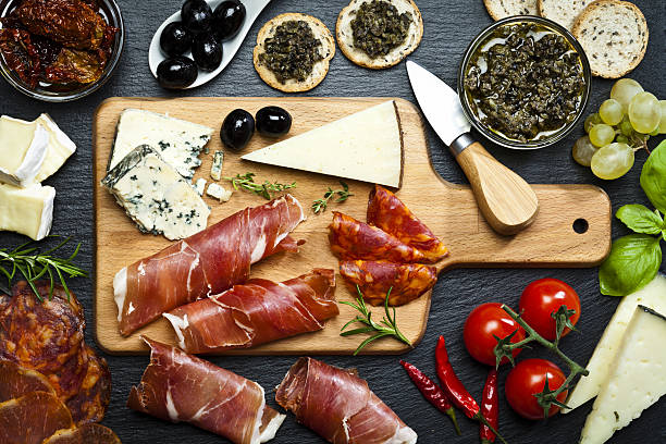 Delicious appetizer on dark slate table Top view of a wooden cutting board on dark slate background filled with a assortment of fine food. The composition includes cold cuts, two glass bowls with dried tomatoes and olives paste, various types of cheese, prosciutto, salami, black olives and some herbs. DSRL studio photo taken with Canon EOS 5D Mk II and Canon EF 100mm f/2.8L Macro IS USM crostini photos stock pictures, royalty-free photos & images