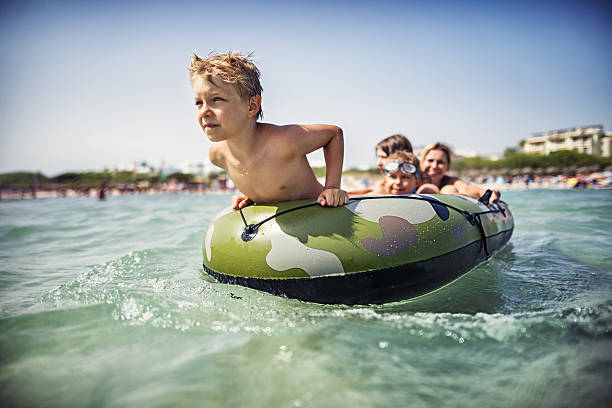 Family is playing with inflatable boat at sea. Three kids sitting in the boat and mother is pushing the boat from behind.