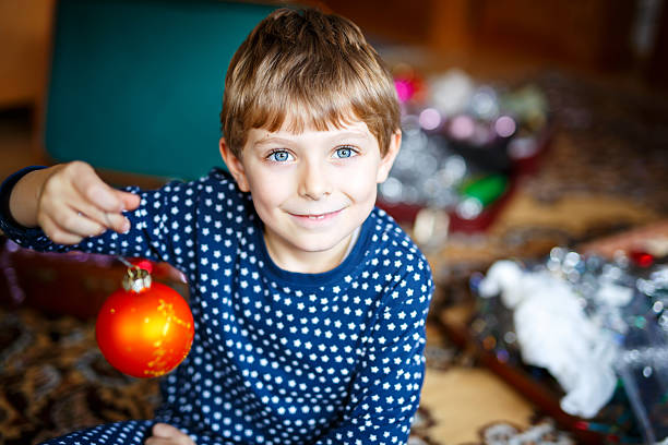 Cute little kid boy decorating Christmas tree with colorful balls and decorations and lights. Happy child having fun before Xmas. Boy in pajama celebrating traditional winter holiday.