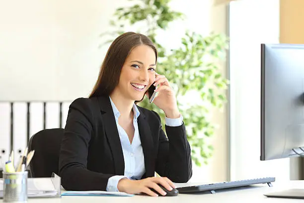 Portrait of a happy executive wearing suit calling customer service on the phone and looking at camera sitting on a desk in the office