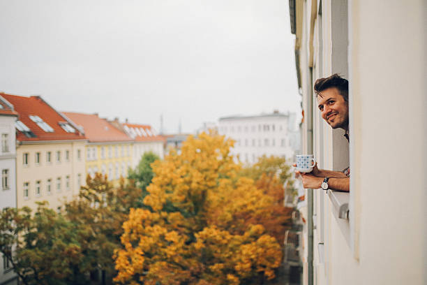 Man looking through the apartment window in Berlin Prenzlauer Berg A young man is drinking coffee and looking from an apartment window, enjoying the afternoon on a cloudy Autumn day in Berlin, Prenzlauer Berg. german people stock pictures, royalty-free photos & images