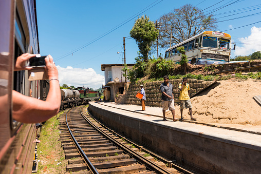Ella, Sri Lanka - March 3, 2015: Old steam train passing the station with some passengers waiting. A tourist is taking photo from the train window. Up the street local bus is parked. Sunny day, blue sky. Scenic view. 