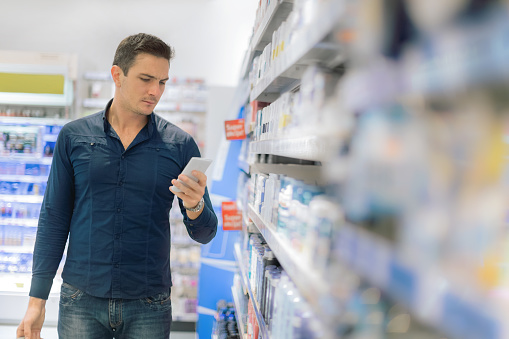 Man using smart phone in local supermarket, standing next to cosmetics