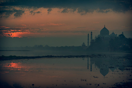 Word famous Taj Mahal in Agra, India - seen from an usual standpoint at the banks of Yamua River. Early morning on a misty day, with reflections in the River. 