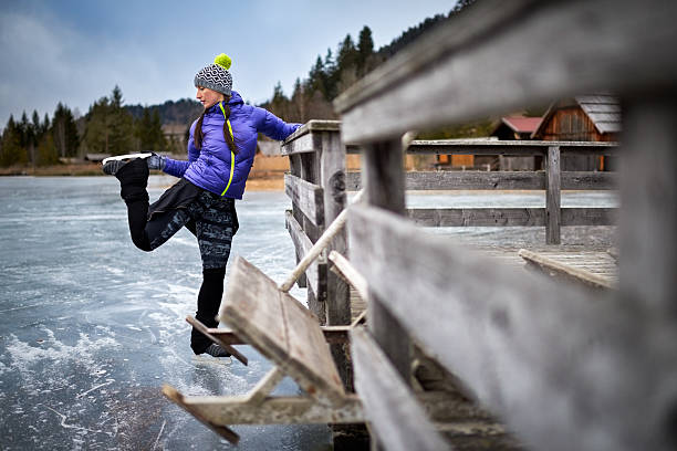 Woman with winter clothes holding the wooden fence on the frozen lake and looking at ice skate shoe. Cabins and hills are in the background.