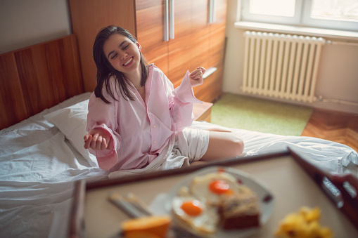Beautiful young woman in pajamas sitting in her bed and smiling, surprised with breakfast in bed
