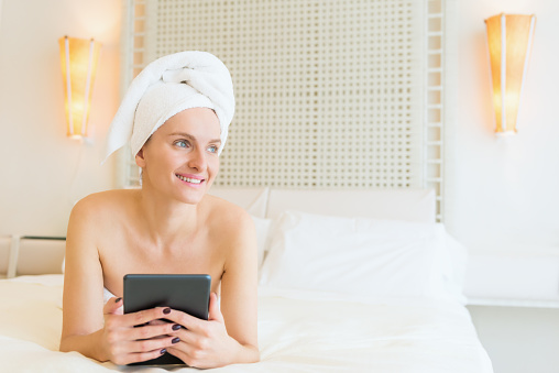 Horizontal color image of beautiful young woman wrapped in towel relaxing and lying on bed after spa beauty treatment. Cheerful woman feeling comfortably, enjoying splendid moment, sincerely smiling and using digital tablet.