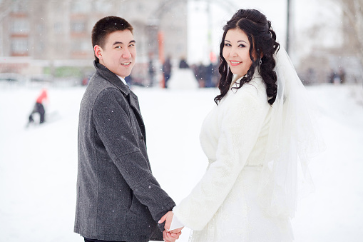 Winter wedding, bride and groom holding hands looking at the camera, classic portrait of couples in the snowy street, frost. Asian people.