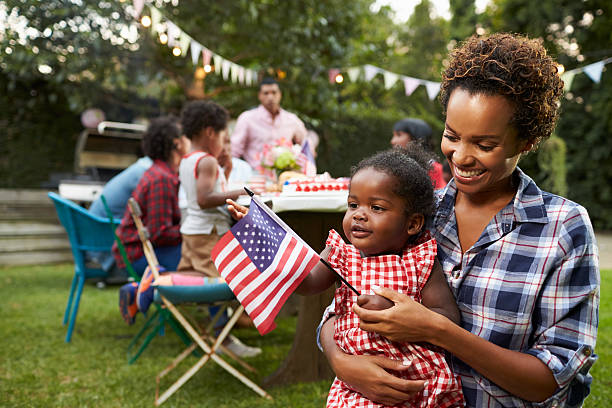 madre negra y bebé sosteniendo la bandera en el jardín del 4 de julio - fourth of july family flag american flag fotografías e imágenes de stock