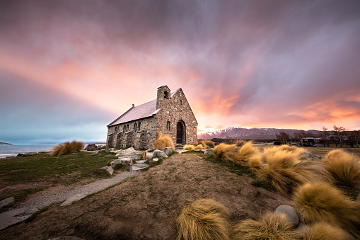 Church of the Good Shepherd on the shores of Lake Tekapo on the South Island of New Zealand. A fiery sunrise throws beams of light out from behind the church making it look like the light is emanating from the church itself. A strong wind blows around the tussocks leading to the church.