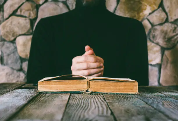 male in black clothes in front of an old open book. the concept of prayer, study, witchcraft. selective focus