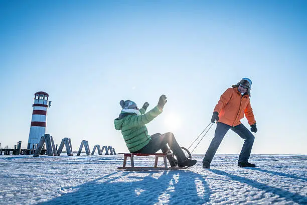 active senior couple man pulling woman on toboggan on frozen lake neusiedl in austria cold sunny winters day backlight