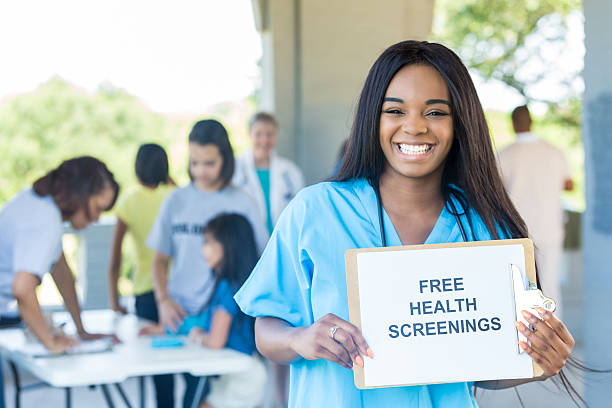 Cheerful young African American nurse promotes health fair Confident African American female nurse holds 'Free Health Screenings' sign to promote health expo. Patients and doctors are in the background. exhibition stock pictures, royalty-free photos & images