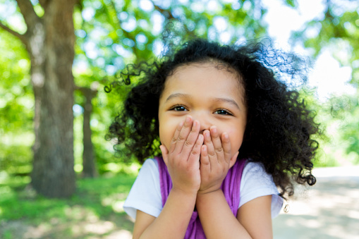 Beautiful preschool age girl covers her mouth in excitement while walking on a footpath in the park.