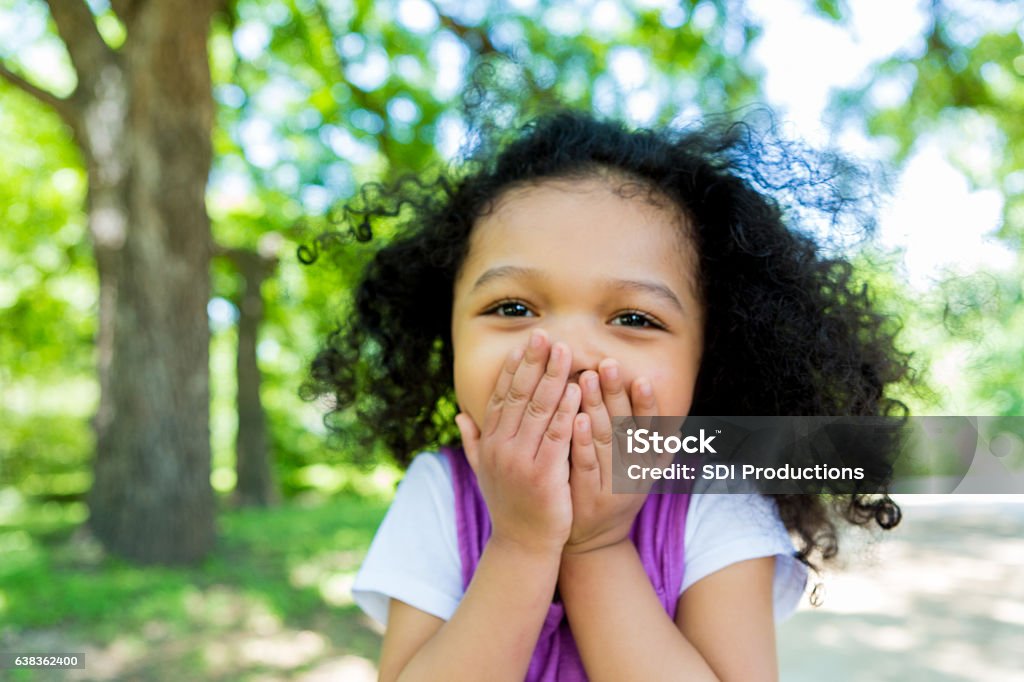 Adorable mixed race girl in the park Beautiful preschool age girl covers her mouth in excitement while walking on a footpath in the park. Child Stock Photo