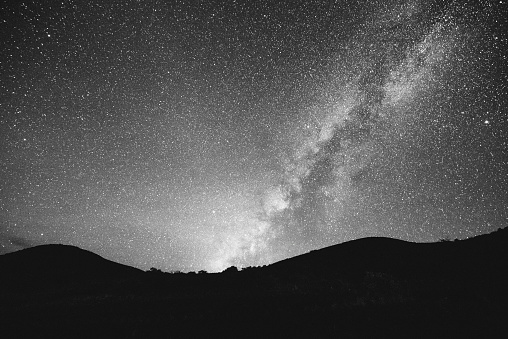 This is a horizontal, black and white, royalty free stock photograph of a  multicolored starry night sky with the Milky Way. The edge of the mountains visible at a high elevation at Mauna Kea Kona, Hawaii are silhouetted. The night sky is multicolored gradient filled with stars. Photographed with a Nikon D800 DSLR camera.