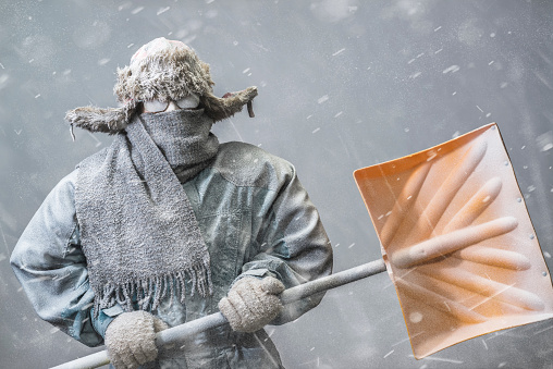 A determined frozen Caucasian male looking at the camera, all bundled up in a fur trappers hat, parka, scarf and gloves, wearing glasses and carrying a yellow snow shovel, covered in snow and frost from the driving snow of a blizzard, heads out to shovel snow.