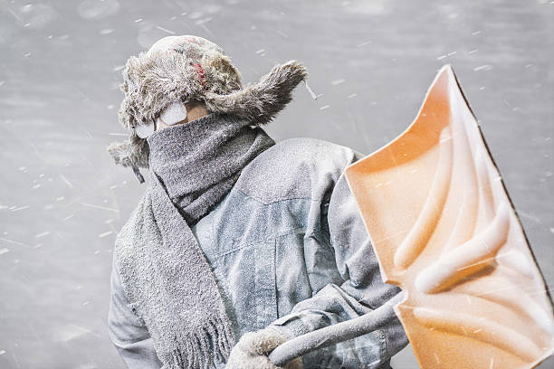A determined frozen Caucasian male, all bundled up in a fur trappers hat, parka, scarf and gloves, wearing glasses and carrying a yellow snow shovel, covered in snow and frost from the driving snow of a blizzard, heads out to shovel snow.