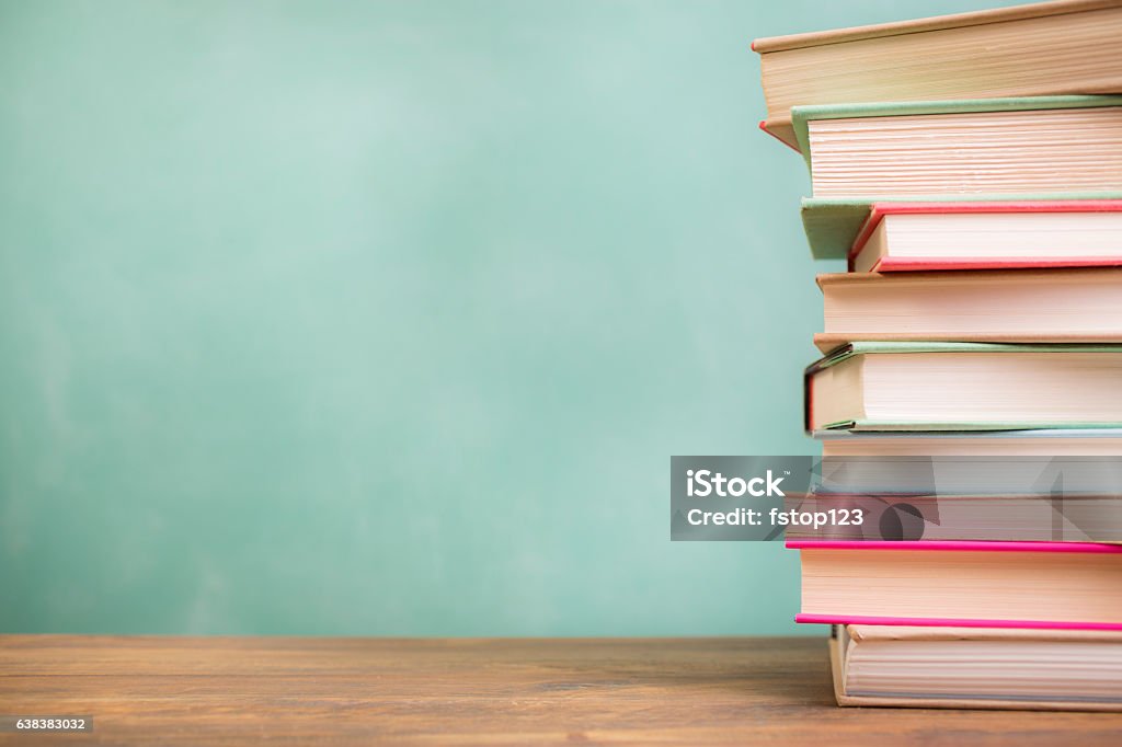 Textbooks stacked on school desk with chalkboard background. It's back to school time!  A large stack of textbooks to side makes frame composition.  The pile of objects lies on top of a wooden school desk with a green chalkboard in the background.  The blank blackboard in the background makes perfect copyspace!  Education background themes. Book Stock Photo