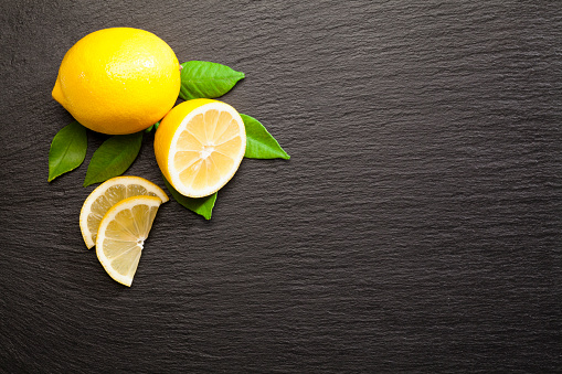 Lemon fruit on black slate background shot directly above. A whole lemon comes from the upper left corner of the frame while one fruit is cut in slices. There is a copy space at the center. Low key DSRL studio photo taken with Canon EOS 5D Mk II and Canon EF 100mm f/2.8L Macro IS USM