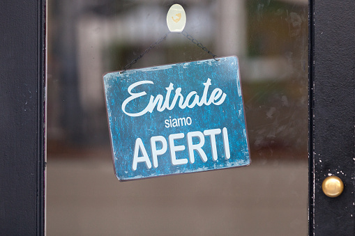 Sign attached to a glass door saying in Italian “Entrate, siamo aperti”, meaning in english “Come in, we’re open”.