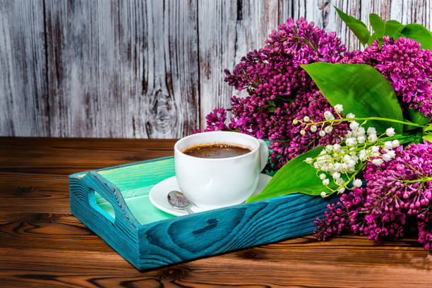 composition with aroma cup of coffee on tray and bouquet beautiful spring lilac, lilies of the valley flowers on brown wooden table, close up
