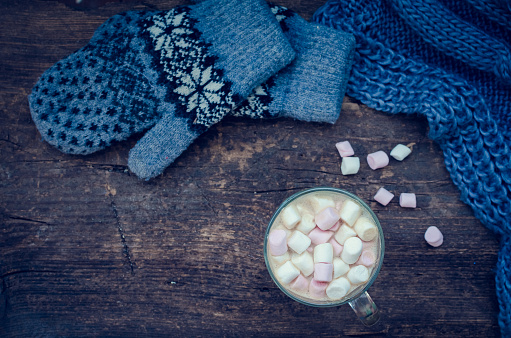 Cozy winter home background, glass of hot cocoa with marshmallow, warm knitted scarf and mittens on an old wooden board background with space for text. Christmas drink. Top view. Copy space.