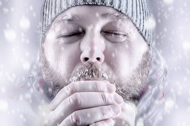 Man freezing in snow storm white out close up Freezing cold man in snow storm white out trying to keep warm by blowing into his hands. Wearing a beanie hat with eyes closed and winter coat. Frost and ice on his beard and eyebrows. frost face stock pictures, royalty-free photos & images