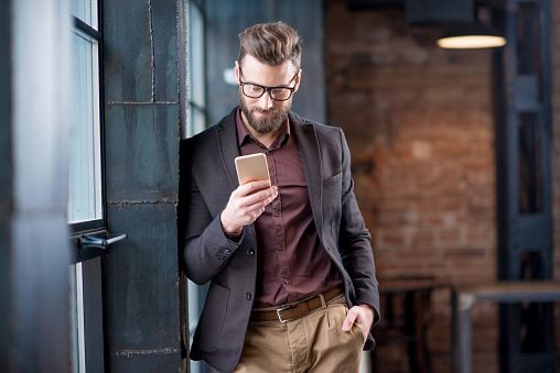 Handsome caucasian businessman dressed in the suit reading with his smart phone near the window in the loft interior studio