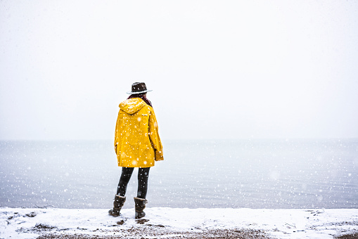 Young woman wearing yellow raincoat and cowboy hat standing by the sea under snow.