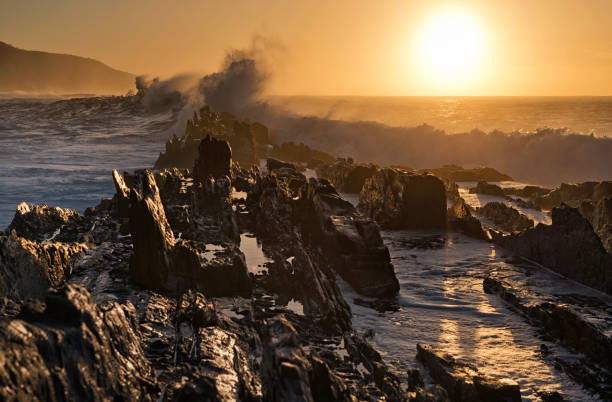 Waves crash against jagged rocks during sunset at Storm's River, South Africa