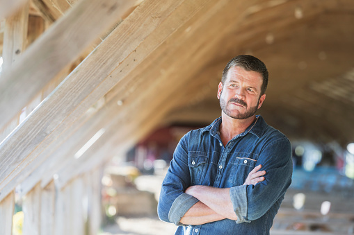 Mature man standing in a barn, serious but confident, with his arms folded, looking away. He is a farmer, owner of a small agricultural business.