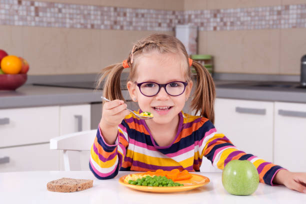 Happy child girl eating vegetables and laughs Happy child girl with glasses eating vegetables and laughs in a kitchen happy green peas stock pictures, royalty-free photos & images
