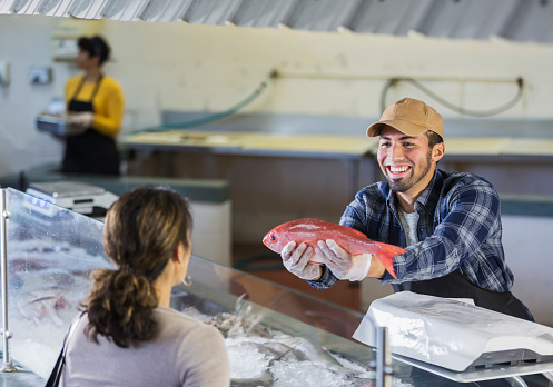 An hispanic man working in a seafood market helping a female customer who wants to buy a fish. He is showing her a whole red snapper before he weighs it on a scale.