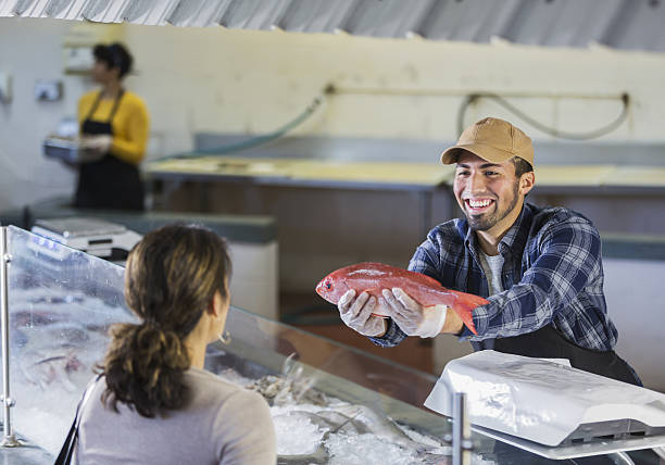 Worker in seafood market helping customer An hispanic man working in a seafood market helping a female customer who wants to buy a fish. He is showing her a whole red snapper before he weighs it on a scale. fish market photos stock pictures, royalty-free photos & images